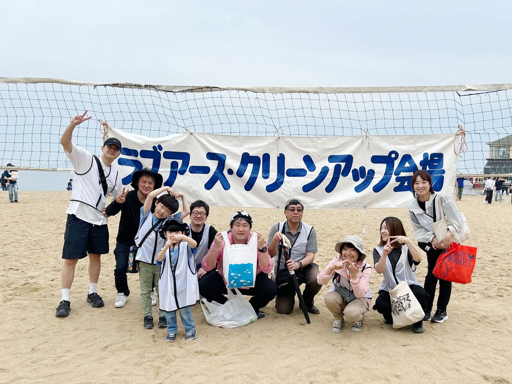 写真：「ラブアース・クリーンアップ会場」の横幕の前で、大人から子どもまで10名ほどの参加者が笑顔でカメラに向かっている集合写真