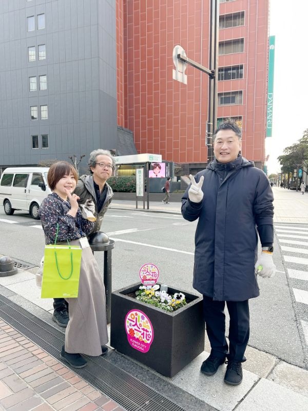 写真：福岡市中央区の渡辺通り沿いに設置された花壇を囲んで、カメラに向かってピースサインする社員3名。後方に福岡大丸の看板が見える