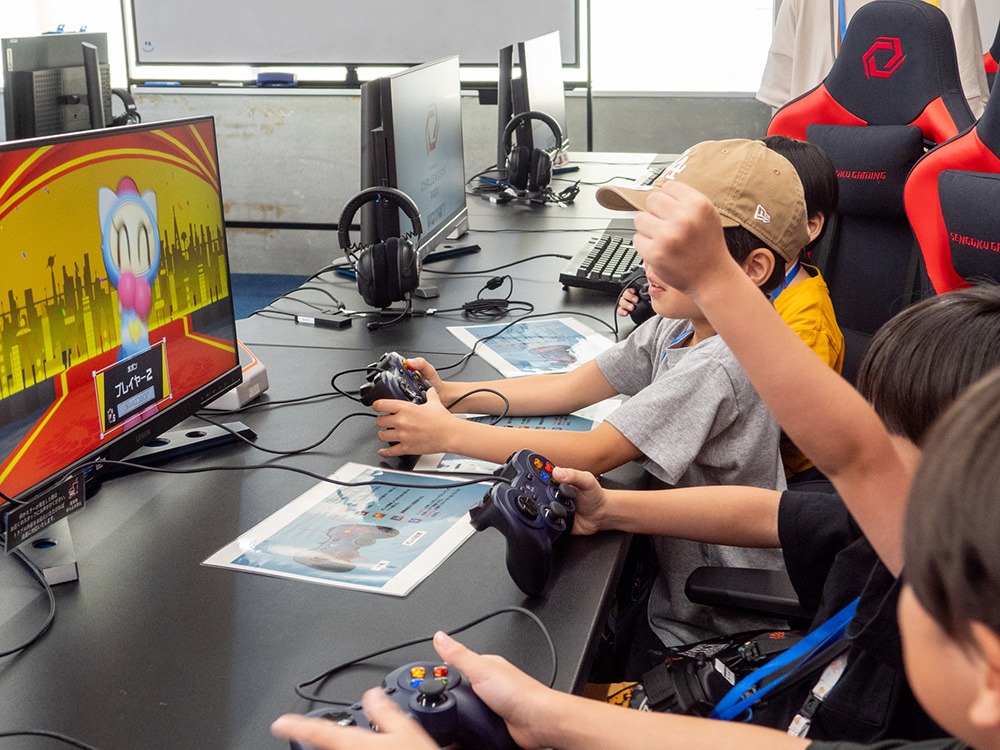 写真：モニターを置いたデスクに子どもが4人並んで座り、それぞれコントローラーを持ってゲームしているようす