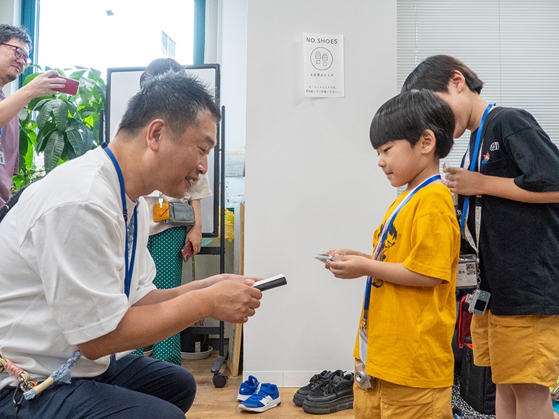写真：男性と男の子が名刺交換しているようす。男性はしゃがんで男の子の目線に高さをあわせている。男の子のうしろにもう一人、男の子がいて、話しかけている