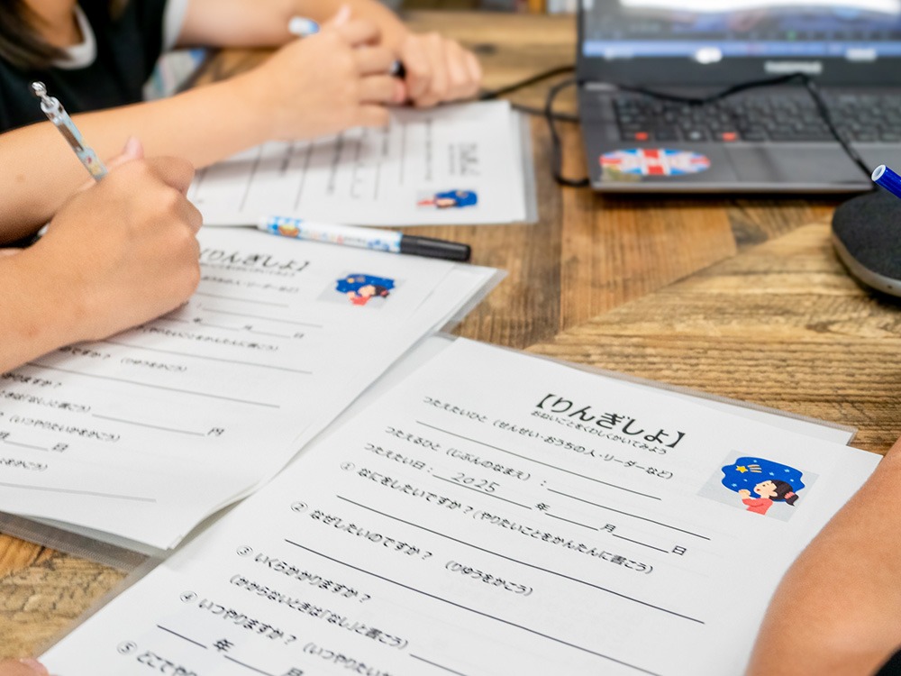 写真：テーブルの上に子ども向けに作られた稟議書の教材があり、子どもの手が文字を書き込んでいるようす