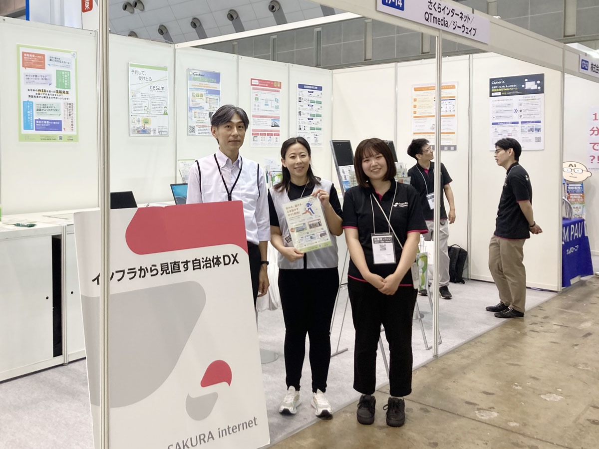 写真：さくらインターネット、ジーウェイブ、QTmediaの共同ブース前で、スタッフ3名が並んでカメラに笑顔を向けている様子