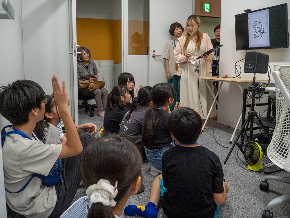 写真：壁に取り付けたモニターの前に女性が立って説明している。子どもたちがフロアに座って説明を聞いており、挙手している子どももいる。フロアの奥には年配の女性が椅子に座ってモニターを見ている